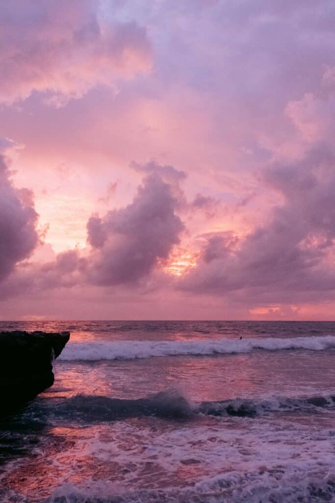 Image d'une mer tranquille depuis la plage, avec un gros rocher sur la gauche et un ciel rose orange et nuageux