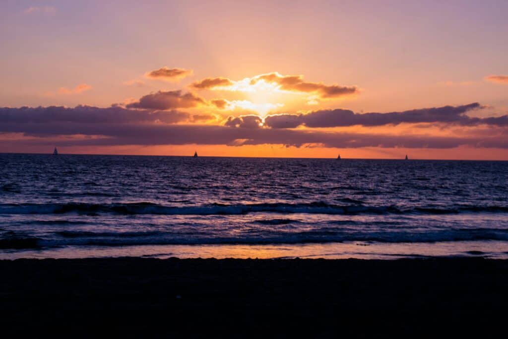 Image d'une mer tranquille depuis la plage, avec quelques bateaux, le soleil derrière les nuages, teintes roses et orangées